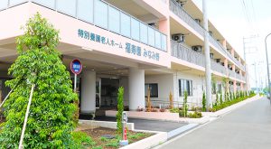 介護施設の建物外観。白い壁面に緑のツタが生い茂り、玄関前に植栽がある。